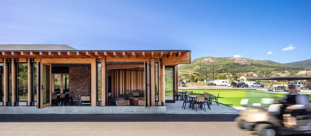 A contemporary low-profile pavilion-style building with extensive floor-to-ceiling glazing, exposed timber roof framing, warm timber posts, and exposed brick walls. The covered outdoor dining terrace is furnished with tables and chairs, opening toward a broad landscaped lawn and a scenic hillside setting. A blurred golf cart passes in the foreground.