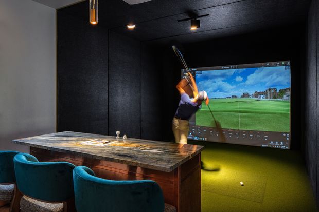A dark, sophisticated golf simulator room with a large projected golf-course screen, turf flooring, and a club swinging in front of the image. A substantial stone-topped bar or counter with timber cabinetry and teal upholstered stools occupies the foreground. Black acoustic wall and ceiling surfaces, recessed spotlights, and pendant lighting create a moody entertainment setting.