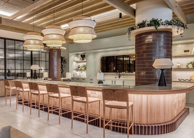 A refined open-plan kitchen and entertaining space dominated by a large curved island with integrated seating for several people. The island has vertical timber-look paneling, a rounded stone or composite benchtop, and a dark tiled plinth. Two cylindrical columns clad in glossy dark brown vertical tiles support a warm timber ceiling structure. Multiple layered pendant lights hang above the island, while a rear kitchen wall includes pale cabinetry, open shelving, a sink and integrated appliances. Black-framed glazed doors or partitions and abundant indoor planting add contrast. The overall interior is warm, tactile and upscale, with a contemporary hospitality-inspired design.
