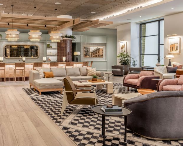A spacious, styled open-plan hospitality or commercial lounge with a bar and service counter at the rear, layered seating areas, and a large patterned area rug. The room combines pale timber flooring, muted sage and cream walls, exposed brick, a decorative timber ceiling feature, pendant lighting, contemporary lounge furniture, and tall black-framed windows. The overall character is warm, refined, and mid-century influenced.
