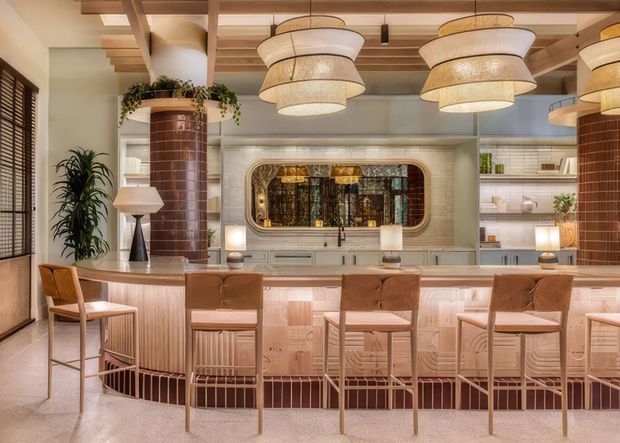 A warm, highly styled home bar or entertaining counter with four upholstered bar stools, a long illuminated curved-front built-in bar, and an extensive backbar. The room features ribbed and tiled joinery, a large decorative mirrored or glazed inset, timber ceiling beams and slats, terrazzo-like flooring, and oversized woven pendant lights. Indoor plants and sculptural lamps add a refined, retro-inspired atmosphere.