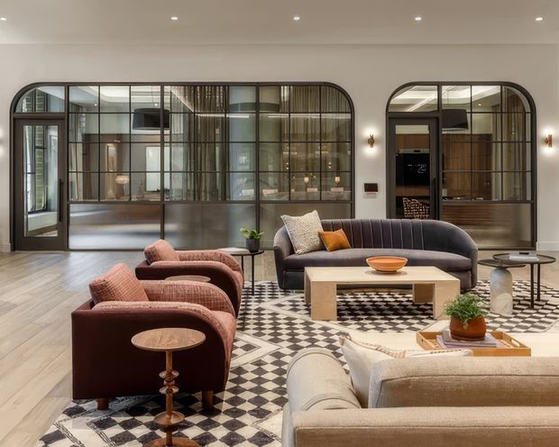 A refined contemporary living room with a layered seating arrangement, featuring a curved dark sofa, rust-coloured lounge chairs, and a large light sofa in the foreground. A substantial pale timber coffee table sits on a bold black-and-white geometric rug, with small round side tables and restrained decorative objects. The room has pale timber flooring, a light neutral ceiling and walls, and striking dark-framed, arched glazed steel-style partitions behind the seating that divide the living area from adjoining spaces. Recessed ceiling lights and warm wall sconces create a polished, welcoming atmosphere.