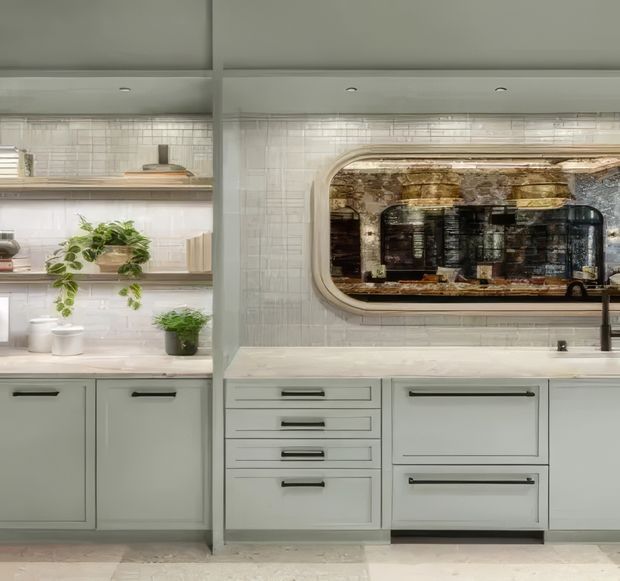 A softly styled contemporary kitchen with pale sage-green cabinetry, white countertops, and a white glazed tile backsplash. Open shelving displays crockery and potted trailing plants, while a large rounded rectangular mirrored or reflective feature inset above the counter adds visual depth. The cabinetry includes drawers and integrated storage, with dark slim pulls and a black tap providing contrast.