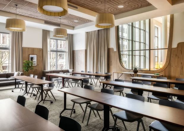A bright, spacious commercial dining or lounge area with multiple long timber tables and black chairs arranged in rows. The room features tall industrial-style gridded windows, full-height drapery, warm timber wall panelling with sculptural curved detailing, patterned acoustic ceiling panels, and large cylindrical pendant lights. A small lounge seating area and indoor plant sit near the windows.