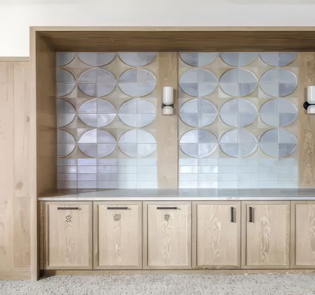 A built-in kitchen or bar-style cabinet wall with pale timber cabinetry, a broad stainless-steel benchtop, and an unusual decorative tiled splashback made from overlapping circular and curved forms in soft white, beige, and pale blue. The cabinetry has dark metal pulls and distressed, handcrafted-looking fronts. A terrazzo-like floor is visible at the bottom.