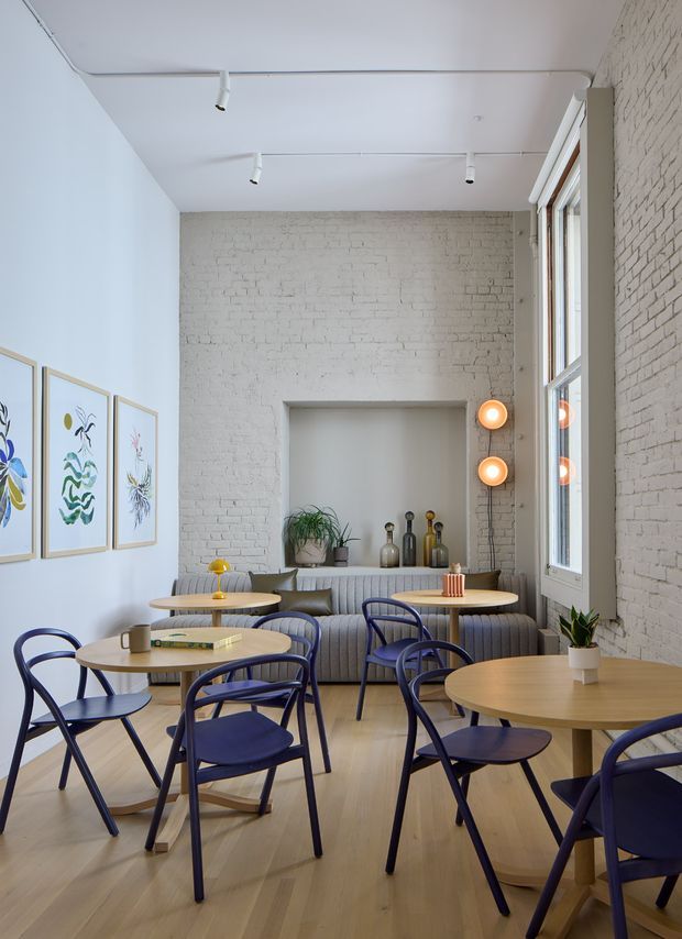 A bright, narrow café or casual commercial dining space with white-painted exposed brick walls, pale timber flooring, and high ceilings. Round light-wood tables are paired with dark blue bentwood-style chairs, while a long ribbed upholstered banquette runs along the rear wall. Track lighting and warm globe wall lights illuminate the room, with framed botanical artwork, small plants, and decorative bottles adding detail. Tall windows line one side, bringing in abundant natural light.