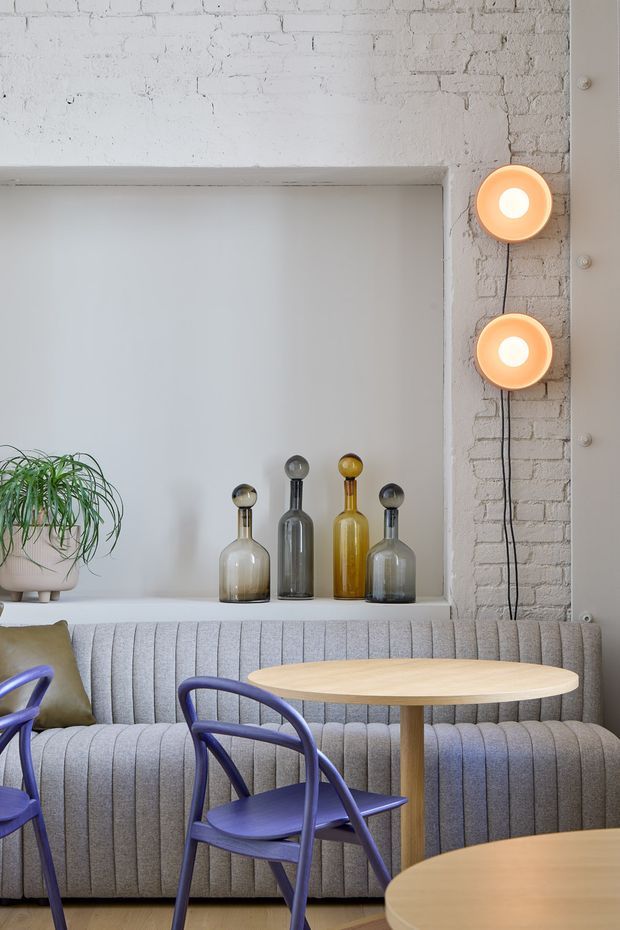 A stylish café or hospitality seating area with a curved, vertically channelled grey upholstered banquette beneath a deep wall niche. The niche contains a potted trailing plant and four decorative glass bottles. A white-painted exposed brick wall frames the space, with two warm globe wall lights and a vertical structural or trim panel. Round light-wood tables and sculptural purple-blue chairs complete the contemporary interior.