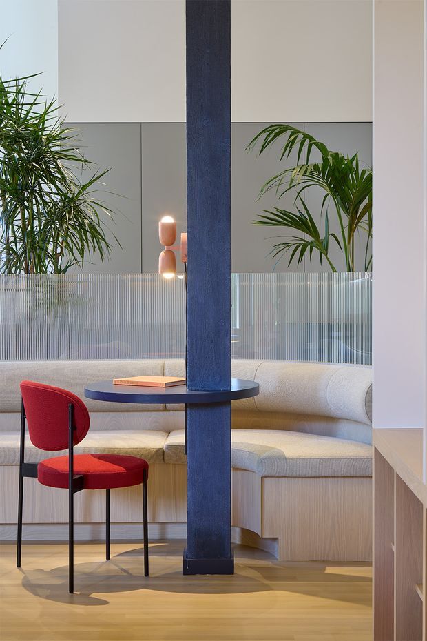 A refined contemporary interior seating area with a curved cream upholstered banquette wrapping around the back and side walls. A deep blue structural column with a small integrated circular shelf divides the composition, while a bright red dining chair provides contrast in the foreground. Pale timber flooring and timber cabinetry frame the space, with indoor palms, a ribbed translucent glass partition, and warm sculptural wall lighting adding texture and softness.