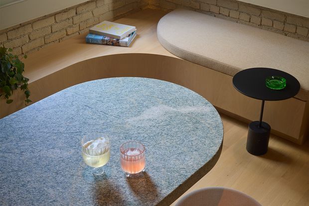 A warm, intimate contemporary living area with a curved built-in timber banquette and pale upholstered seat set against exposed light brick walls. A large rounded coffee table with a blue textured surface dominates the foreground, accompanied by a small black side table and soft upholstered seating. Books, drinks, and a plant add lived-in detail.