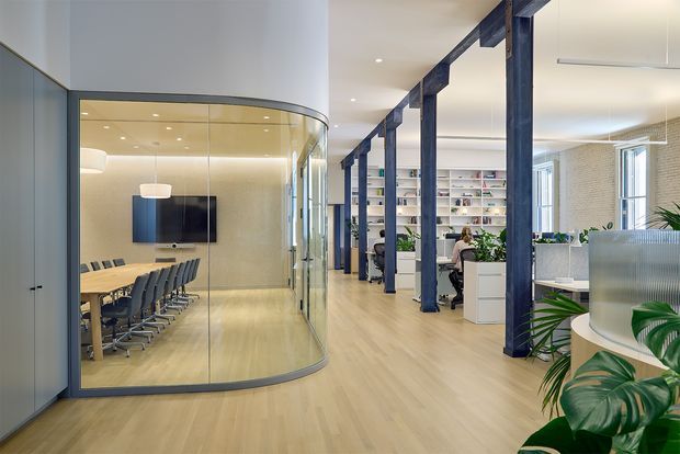 A bright contemporary office interior with an open-plan workspace and a glazed, curved meeting room. The meeting room contains a long wood-toned conference table, rows of dark chairs, a wall-mounted display, and suspended pendant lighting. Beyond it are workstations, white built-in shelving, desks, indoor plants, and exposed dark structural columns and beams. Light timber flooring, extensive internal glazing, and a neutral palette create an airy professional environment.