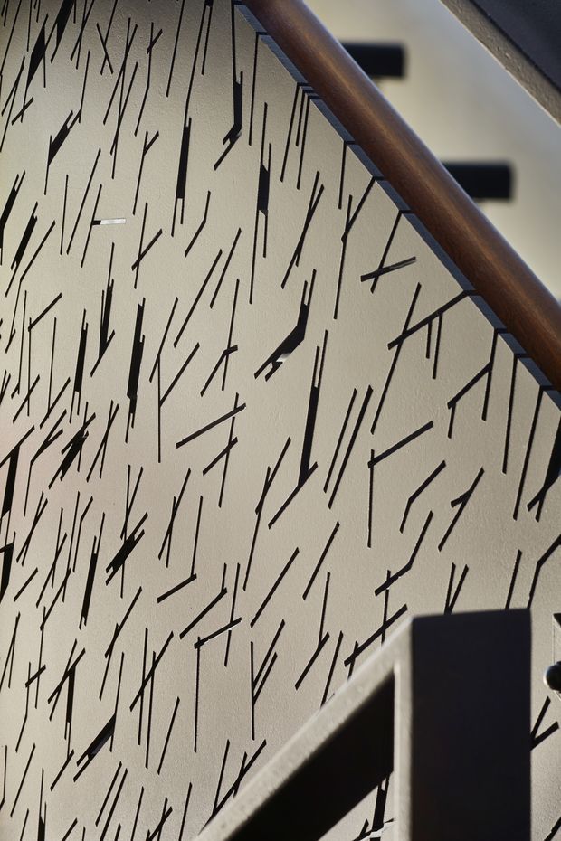 Close-up architectural detail of a stairway with a cream textured wall covered in an irregular black linear pattern. A dark timber handrail runs diagonally across the upper portion, with dark metal balusters and stair elements creating strong geometric shadows. The image focuses on the stair balustrade, wall treatment, and contrast between natural timber and black metal.