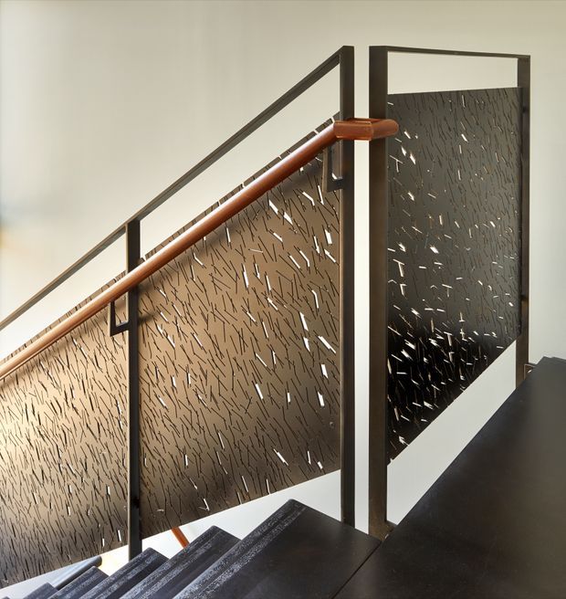 Close-up architectural view of an interior stairway with dark stair treads, slim dark metal framing, a warm timber handrail, and perforated metal privacy balustrade panels. The stair appears contemporary and sculptural, with light filtering through the irregular slotted screens.