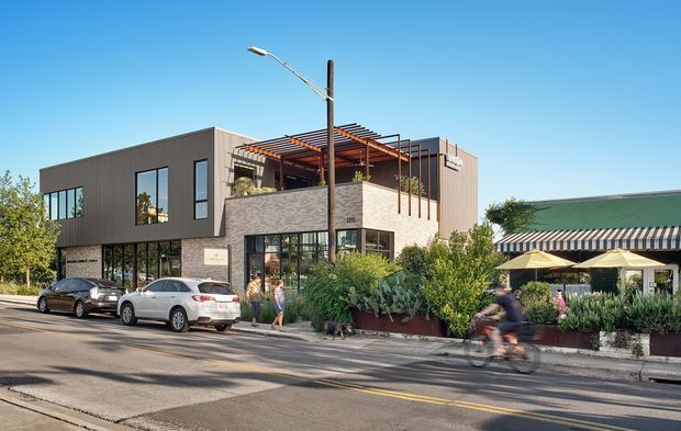 Street-level exterior view of a contemporary multi-storey commercial or mixed-use building with dark vertical cladding, extensive ground-floor glazing, a light masonry-clad upper terrace, and a timber pergola with rooftop planting. The building fronts an urban street with parked cars, cyclists, landscaped planters, and an adjacent low-rise building with a striped awning.