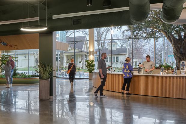 A spacious contemporary commercial lobby or café reception area with a long timber-fronted service counter, polished concrete floor, exposed dark ceiling services and ductwork, pendant lighting, large glazed openings, and indoor planting. People are gathered at and walking through the space. Residential-style buildings and trees are visible outside through the glazing.