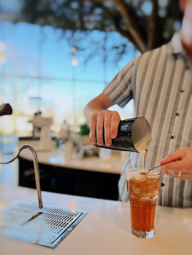 Close-up view of a person preparing an iced beverage at a sleek kitchen or café-style counter. A curved stainless-steel tap and integrated drain grate sit in the foreground, with a pale stone countertop and dark lower cabinetry. Large glazing in the background creates a bright, airy setting, while the person pours a creamy topping into a tall glass of iced tea or coffee.