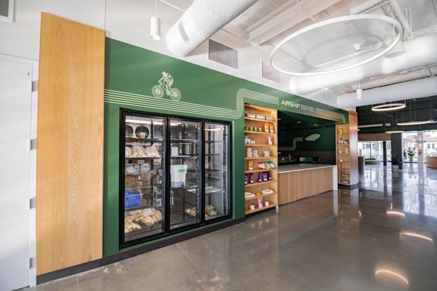 A bright, modern commercial retail interior featuring a green branded service counter and integrated shelving, black-framed refrigerated display cases, timber wall panelling and cabinetry, polished concrete flooring, and an exposed white services ceiling with large circular pendant lighting. Glazed storefront openings are visible at the far end.
