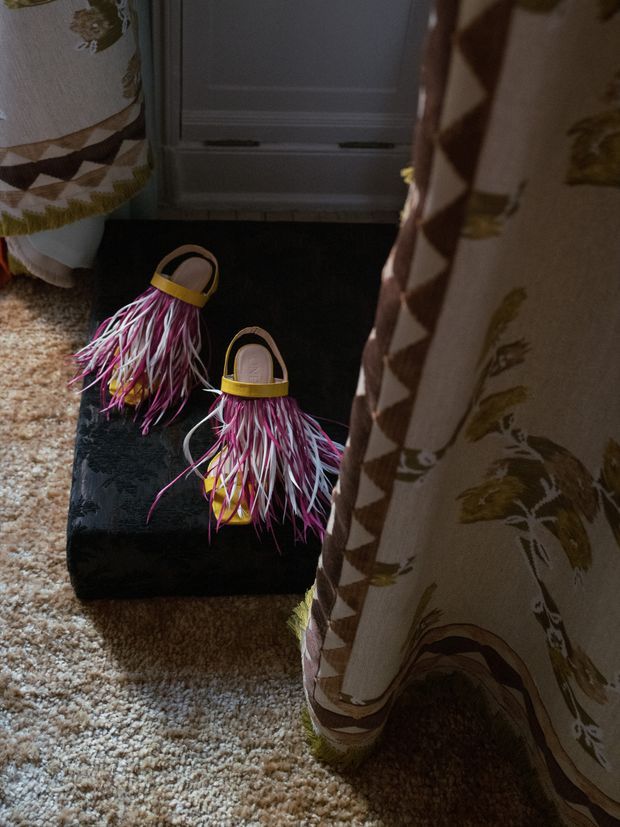 A close-up interior detail showing a pair of colorful feathered sandals resting on a black bag beside a bed or upholstered furniture. A patterned textile drapes prominently in the foreground, with a textured beige carpet and a grey paneled surface in the background. The image has a warm, intimate, lived-in atmosphere.