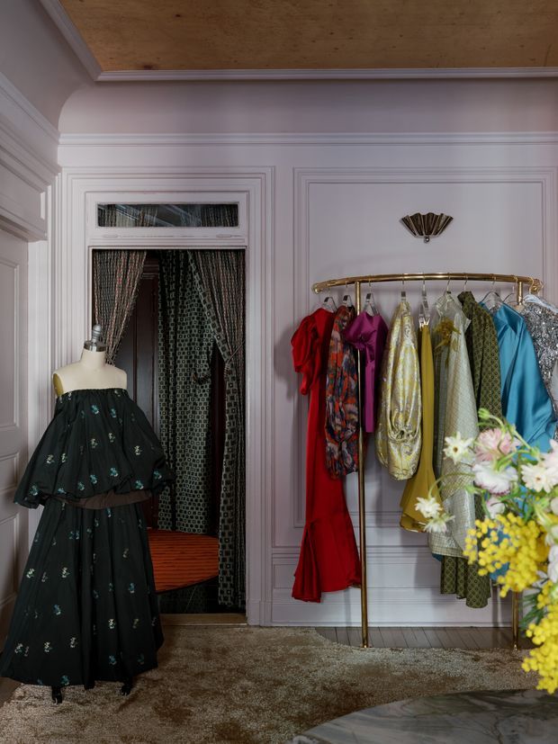 A richly styled dressing room or fashion showroom with ornate pale moulded wall panelling, a warm timber ceiling, and a brass garment rail displaying colourful clothing. A mannequin wears a dark floral dress in the foreground, while a curtained doorway or fitting area and a textured carpet complete the scene. A flower arrangement partially occupies the lower right corner.