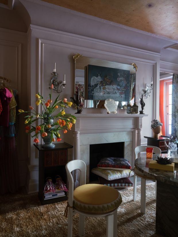 A richly decorated traditional living room with a carved white fireplace mantel, marble surround, ornate wall panelling, and a large gilt-framed mirror. The room contains vintage-style seating, a round table, a patterned rug, decorative objects, floral arrangements, and candle sconces. Warm daylight enters through the window at right, creating an eclectic, historic and layered interior.