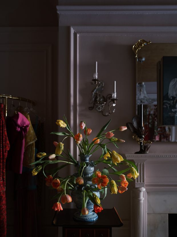 A dark, richly styled traditional interior vignette featuring a large ornate fireplace mantel with decorative moulding, wall-mounted candle sconces, a mirror, and a dramatic arrangement of orange, yellow, pink and red flowers in a blue-and-white ceramic vase. A clothing rack is partially visible at left, while the room is finished in deep mauve-brown tones with low, moody lighting.