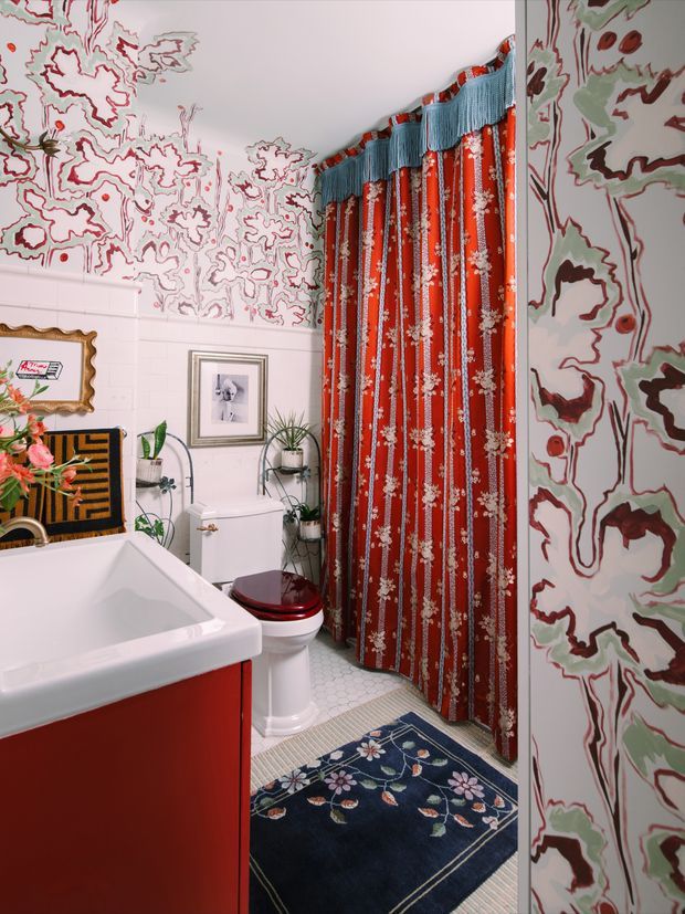 A compact, highly decorative bathroom with a white bathtub and red vanity, a white toilet, patterned floral shower curtain, and bold marbled floral wallpaper wrapping the upper and side walls. The room includes a dark floral bath mat, potted plants, framed artwork, a vintage-style mirror, and layered red, blue, white, and green detailing. The overall look is colourful, eclectic, and maximalist.