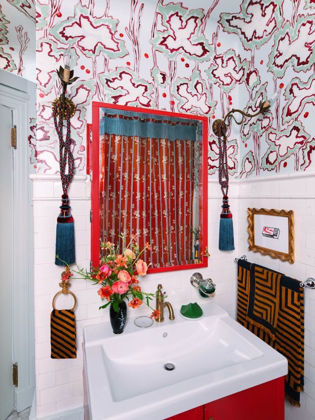 A compact, highly decorated bathroom vanity area with a white rectangular basin set into bright red cabinetry. The walls combine white subway tile wainscoting with vivid abstract red, teal, black and white patterned wallpaper above. A red-framed mirror, ornate tassel wall sconces, patterned black-and-orange towels, brass-toned tapware, framed artwork and a colourful floral arrangement create an eclectic, theatrical look.