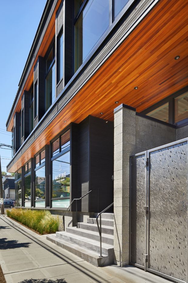 Street-level view of a contemporary multi-storey commercial-style building facade with extensive glazing, dark metal framing and cladding, warm timber soffits and facade accents, concrete block walls, and a textured metal entry gate. Concrete steps with simple black handrails lead to the recessed entrance, while landscaped grasses line the sidewalk.