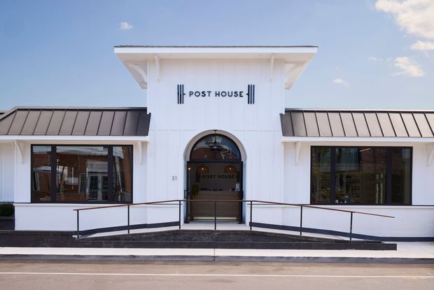 Front exterior view of a low-rise commercial building named Post House. The symmetrical white facade has a projecting central entrance tower with arched glazed doorway, broad eaves, dark standing-seam metal roofs, large dark-framed windows, and an accessible ramp with black metal handrails.