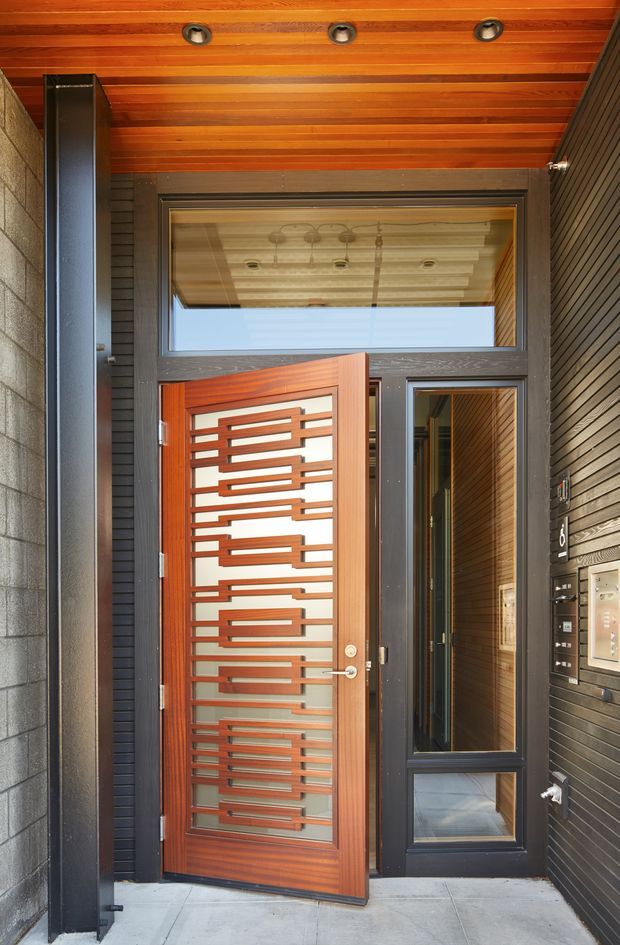 A contemporary residential entryway viewed from outside, featuring a partially open timber front door with a geometric horizontal timber-and-glass insert, dark framed sidelights and transom glazing, dark horizontal wall cladding, and a warm timber-lined soffit with recessed lights. Concrete pavers form the threshold, while visible access and intercom panels are mounted beside the door.