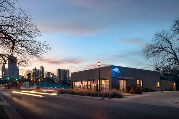 Exterior twilight streetscape showing a low, modern commercial building with a dark box-like facade, extensive floor-to-ceiling glazing, illuminated signage, and a landscaped forecourt. The building sits on an urban street with high-rise buildings visible in the background and vehicle light trails in the foreground.