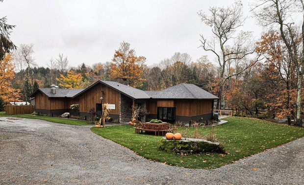 A single-storey dark timber-clad house set within a wooded autumn landscape. The building has multiple pitched roof volumes, a broad gravel driveway, lawn, stone-edged garden beds, and large windows and glazed doors. The scene is photographed from outdoors under an overcast sky.