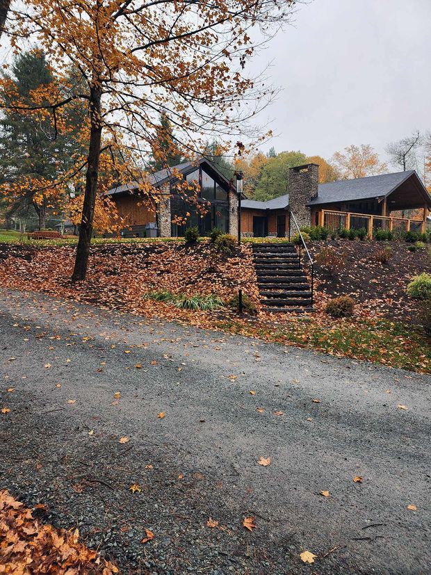 Exterior view of a low-rise, wood-and-stone house set within a mature autumn landscape. The home features several steeply pitched gabled roof forms, dark roofing, timber cladding, substantial rough stone wall sections and a prominent stone chimney. A stepped path leads up from a gravel driveway through landscaped planting toward the covered entrance and glazed central section.