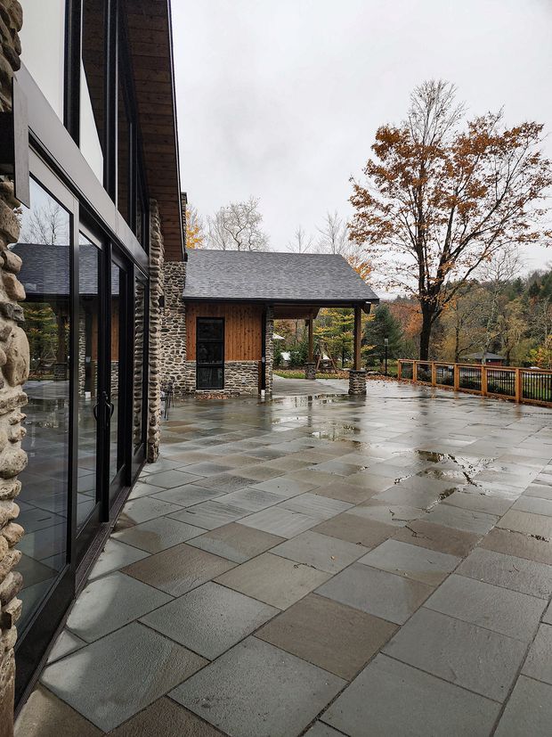 Rainy exterior view of a spacious paved terrace beside a contemporary building. The facade combines extensive black-framed glazing with rough natural-stone walls, while a covered timber-clad pavilion with a dark shingle roof sits beyond the terrace. Stone columns, timber railings, autumn trees and landscaped grounds create a rustic, wooded setting.