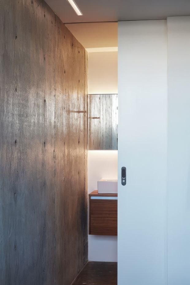 A narrow, minimalist interior passage or entry area viewed through a partially open white sliding door. The space features a full-height textured dark timber wall with visible vertical grain, a dark timber floor, integrated warm strip lighting, and a compact floating timber vanity or cabinet with a white basin at the rear. A reflective surface or opening echoes the timber wall, creating depth. The palette is restrained and contemporary, with warm wood contrasted against white joinery and soft illumination.