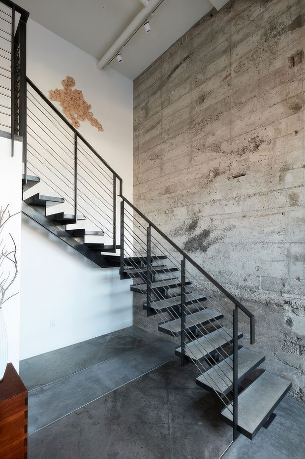 Interior view of a contemporary industrial stairway with an open-riser black metal staircase, slim horizontal cable railings, and a substantial exposed textured concrete wall. The space has polished dark concrete flooring, white walls, exposed ceiling services, track lighting, and a small warm-toned wall artwork.