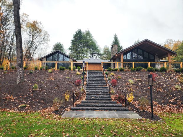 Exterior view of a low, modern timber-and-glass house set on a landscaped, sloping site. A central flight of dark steps rises through garden beds to a timber fence and the home, which has broad glazed gable ends, dark rooflines, and a prominent stone chimney. Mature deciduous and evergreen trees surround the property in an autumn setting.