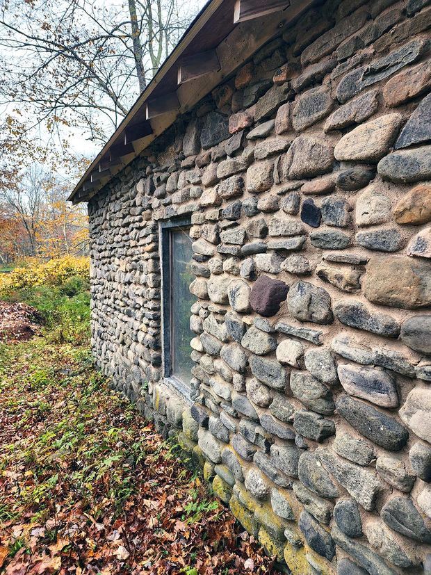 Close exterior view of a low, rustic building with thick walls made from irregular rounded natural stones and visible mortar joints. A narrow dark-framed window is set into the stonework, beneath a simple timber roof overhang with exposed eaves. The building sits in a wooded, leaf-covered landscape during autumn.