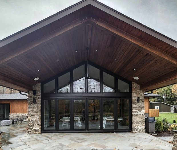 A single-storey gabled pavilion or house extension viewed from the exterior. The facade features extensive dark-framed floor-to-ceiling glazing beneath a large timber-lined pitched gable roof, supported by substantial rough natural-stone piers. A paved terrace sits in front, with a dining area visible through the glass and landscaped grounds around the building.