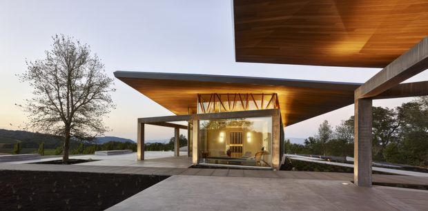 A modern single-storey pavilion-style residence viewed from an expansive concrete terrace. The building features dramatic cantilevered flat roof planes with warm timber soffits, slender dark structural frames, extensive glass walls, and a central glazed living volume. The surrounding setting includes landscaped terraces, mature trees, and distant rolling hills visible beyond the property.