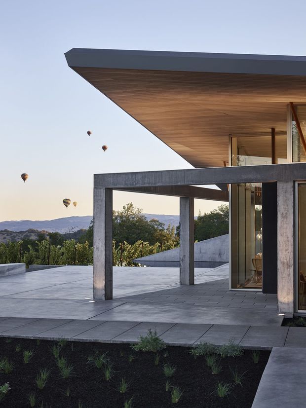 A contemporary flat-roofed house viewed from an expansive outdoor terrace. The structure features a deep cantilevered roof with a warm timber-lined soffit, exposed dark concrete or metal structural framing, extensive floor-to-ceiling glazing, and large grey paved surfaces. Minimal landscaping surrounds the terrace, while hills and hot-air balloons are visible in the distant background.