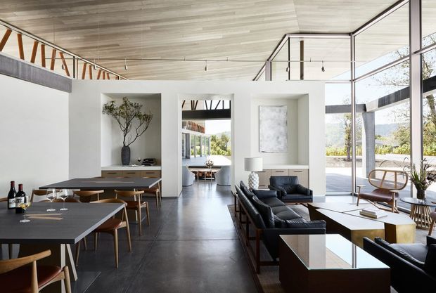 A spacious open-plan contemporary living and dining area with floor-to-ceiling glazing, polished dark flooring, white walls, timber-lined ceiling, and a dramatic double-height volume. The room includes a large dining table, lounge seating, built-in cabinetry, and a sculptural staircase or upper-level balustrade visible at the left. Abundant natural light creates a refined indoor-outdoor connection.