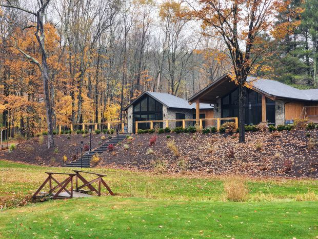 A low, modern house nestled in a wooded autumn landscape. The home features dark timber and masonry walls, extensive dark-framed glazing, multiple gently pitched roof forms, and a raised deck or terrace with timber posts and railings. A landscaped sloping garden with steps, lawn, and woodland surrounds the building.