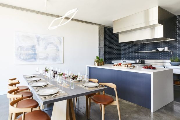 Bright contemporary open-plan kitchen and dining area. A long dining table is set with plates, glassware and floral arrangements, surrounded by timber-framed chairs. The kitchen features a large island with dark blue cabinetry, white benchtops, a dark blue small-format tiled splashback, extensive white cabinetry and a stainless-steel rangehood. Concrete-look flooring and a sculptural pendant light complete the refined, high-contrast interior.