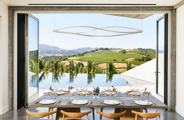 A bright, contemporary dining room opens through fully retracted glazed doors onto a terrace with a reflective pool and vineyard landscape beyond. A long timber dining table is set for a large group with white crockery and glassware, surrounded by sculptural timber dining chairs. The room has white walls, a pale ceiling, a large frameless opening, and a linear sculptural pendant light.