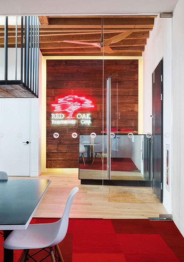 Interior view of a contemporary office or commercial workspace with a glazed meeting room or reception enclosure. The space features warm exposed timber ceiling beams, a dark reclaimed timber feature wall, white walls, glass partitions with metal fittings, light timber flooring, and vivid red carpet accents. A red neon sign, communal table, chairs, and ceiling fan add a distinctive industrial-retro character.