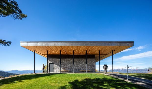 A minimalist pavilion-like building set on a grassy elevated site, with a broad thin flat roof projecting far beyond a compact dark stone-clad enclosure. Slender black columns support the roof, while expansive open sides frame a distant blue landscape and sky. The image is a clear exterior architectural view in bright daylight.