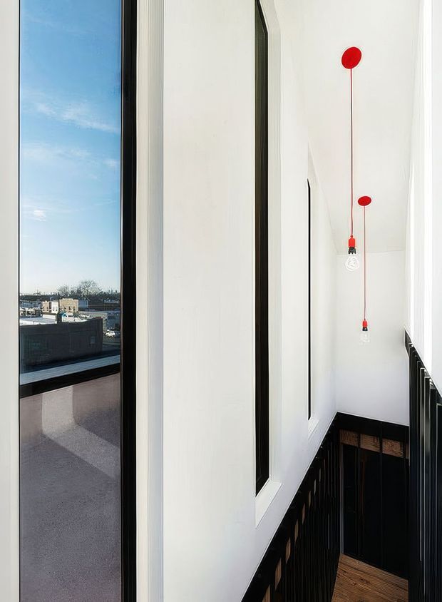 A narrow, bright interior stairway or landing with a dramatic double-height white wall, tall black-framed windows, and black vertical stair balustrades. Red pendant lights hang at different heights, while timber stair treads and a dark timber doorway add warmth. An urban rooftop view is visible through the glazing.