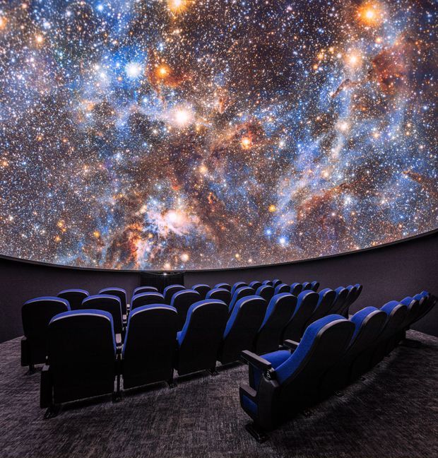 A dark immersive auditorium with rows of upholstered cinema seats facing a large curved dome ceiling displaying a vivid star-field and galaxy projection. Blue-edged black seating, dark walls, and charcoal carpet create a dramatic planetarium or immersive cinema atmosphere.
