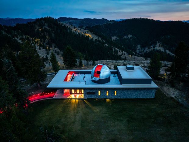 A dramatic aerial dusk view of a low-profile contemporary building set in a mountainous, heavily forested landscape. The structure has broad flat roofs, dark stone or textured exterior walls, illuminated interior glazing, and a prominent rooftop observatory dome with a red-lit opening. A curved illuminated walkway leads through the dark landscape to the building.