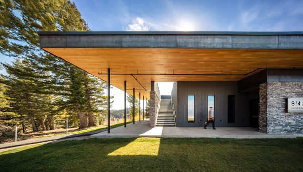 Exterior view of a contemporary low-profile building with a broad flat roof and deep cantilevered timber soffit. Slim black posts support a covered entry walkway, while dark vertical cladding, exposed stone masonry, concrete paving, and a central stair create a strong modern composition. The building is set among mature trees and lawn under a bright blue sky.